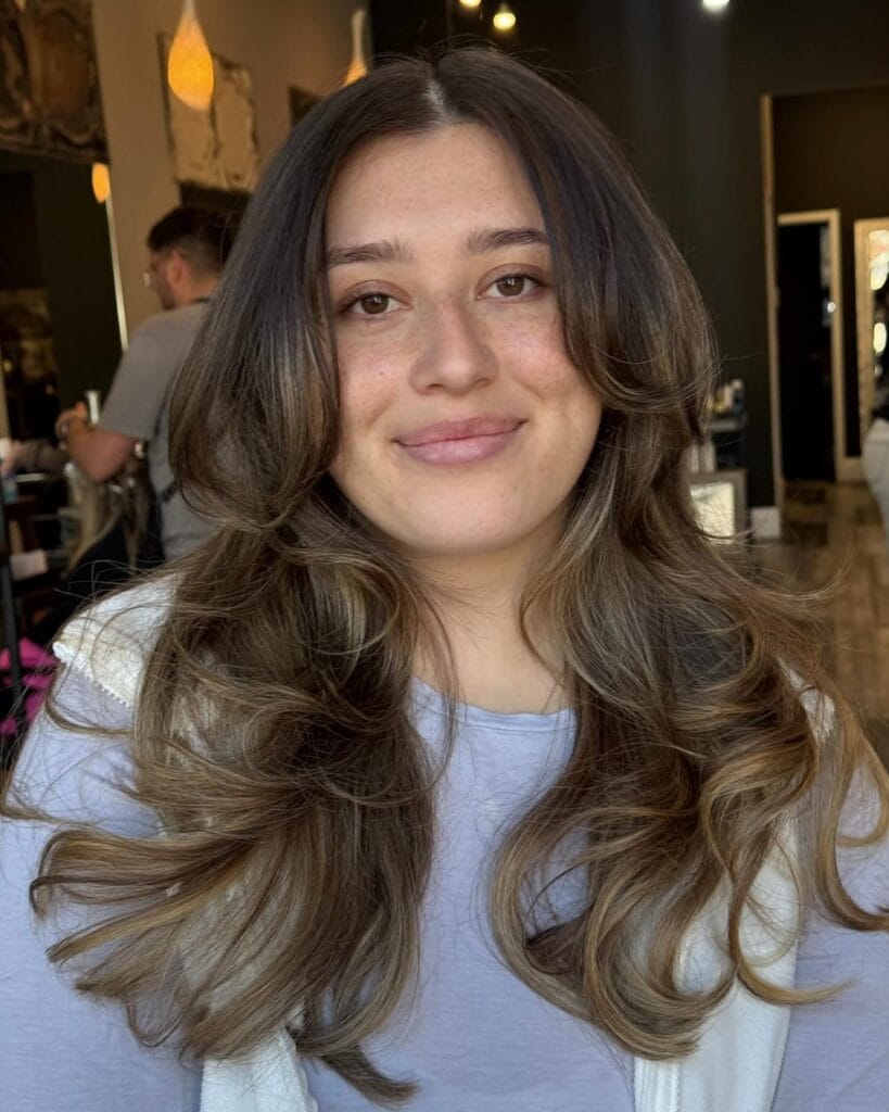 Long layered haircut with soft waves and curtain bangs, featuring blended light brown highlights for added movement and dimension.