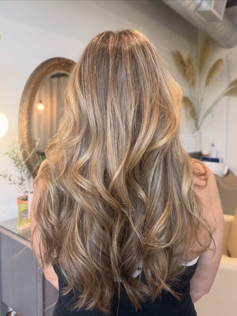 Long layered brunette hair styled in loose waves with soft honey blonde highlights blended throughout for a natural, dimensional finish.
