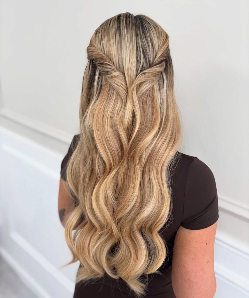 Long layered blonde hair with soft waves styled in a half-up twisted crown that blends into flowing, loose lengths for a soft romantic look.