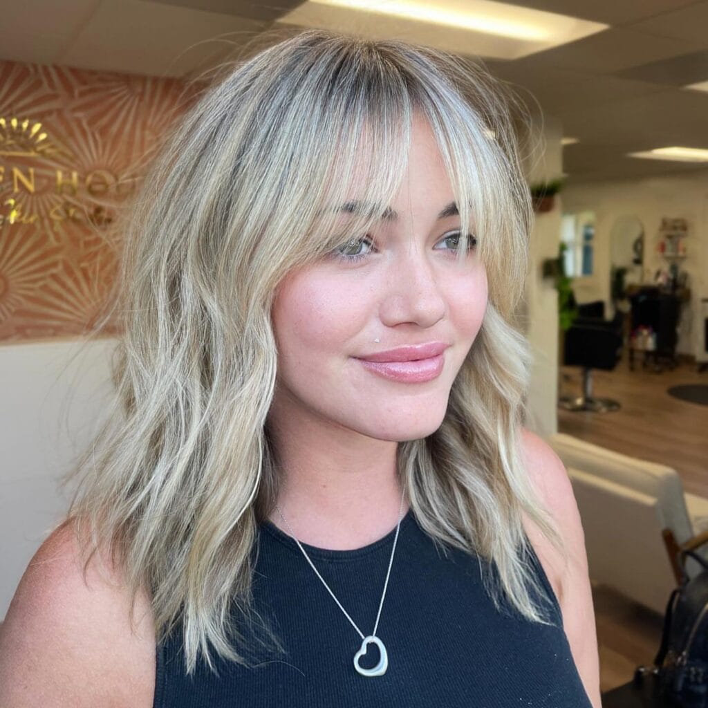 Medium-length blonde shag haircut with soft wavy layers and airy curtain bangs that frame the face for a relaxed, modern textured look.