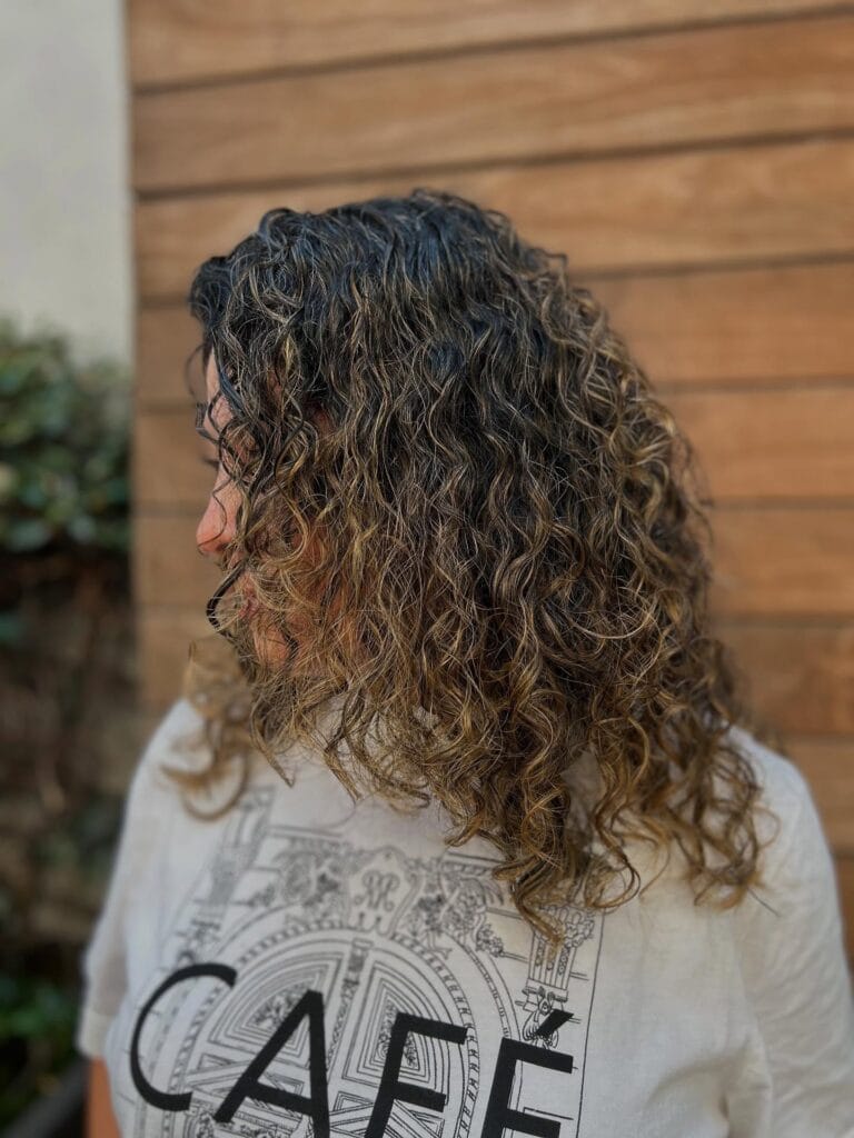 Mid-back length curly hair with long layers and natural volume, featuring defined spirals and subtle caramel highlights for added dimension.