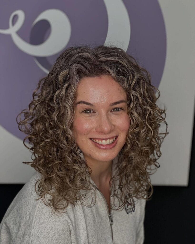 Shoulder-length layered curls with a natural center part and soft gray highlights, creating full, bouncy volume around the sides.