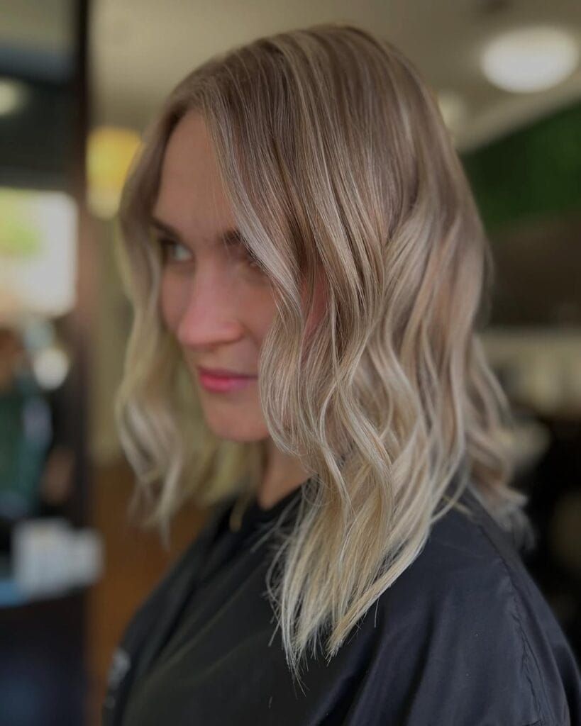Shoulder-length blonde hair with a soft center-to-slightly-off-center part, subtle balayage highlights, and loose natural waves that add light texture through the ends.