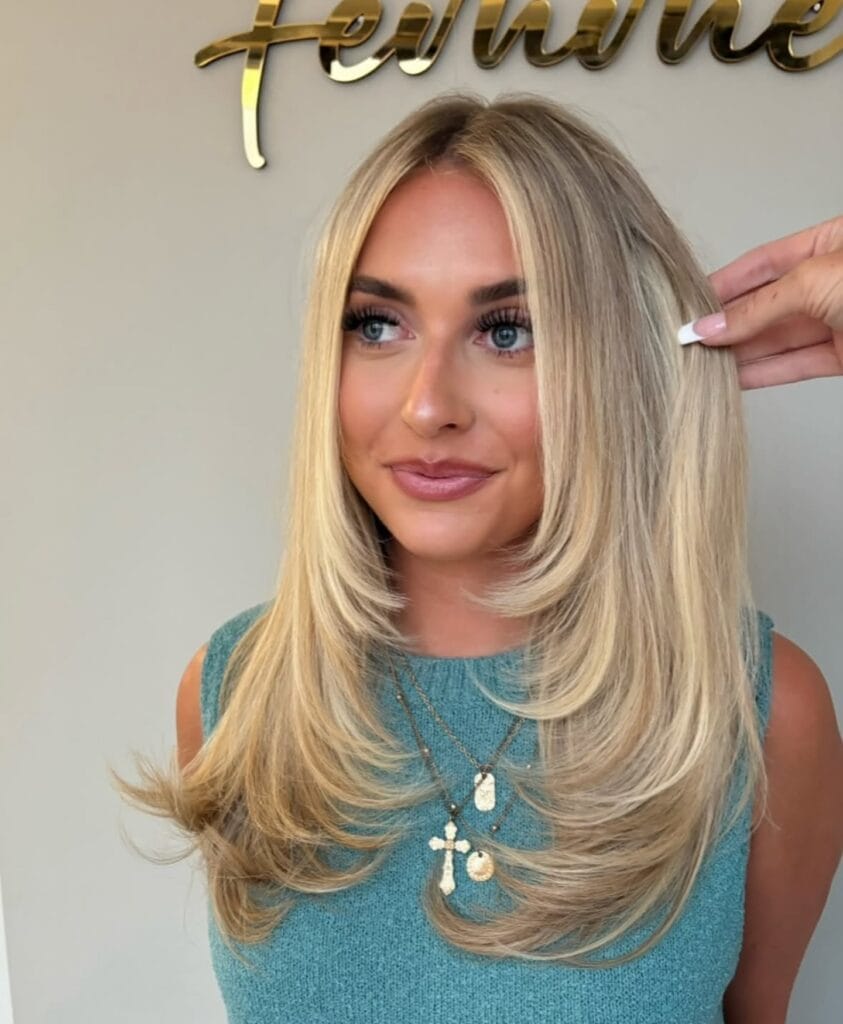 Mid-length blonde hair with a center part, soft face-framing layers, and smooth blowout styling that curves gently inward at the ends for a polished, voluminous finish.