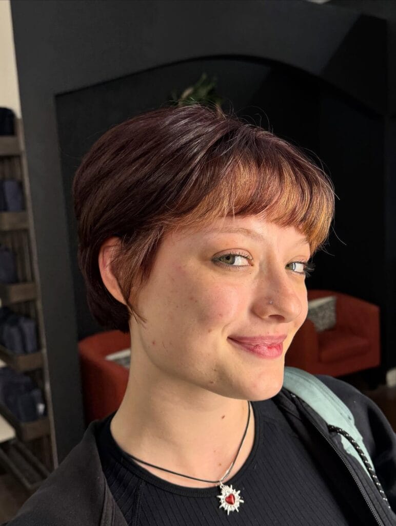 Short layered pixie cut on fine straight hair with soft side-swept bangs, tapered sides and nape, and warm auburn-brown color with subtle lighter copper highlights at the fringe.