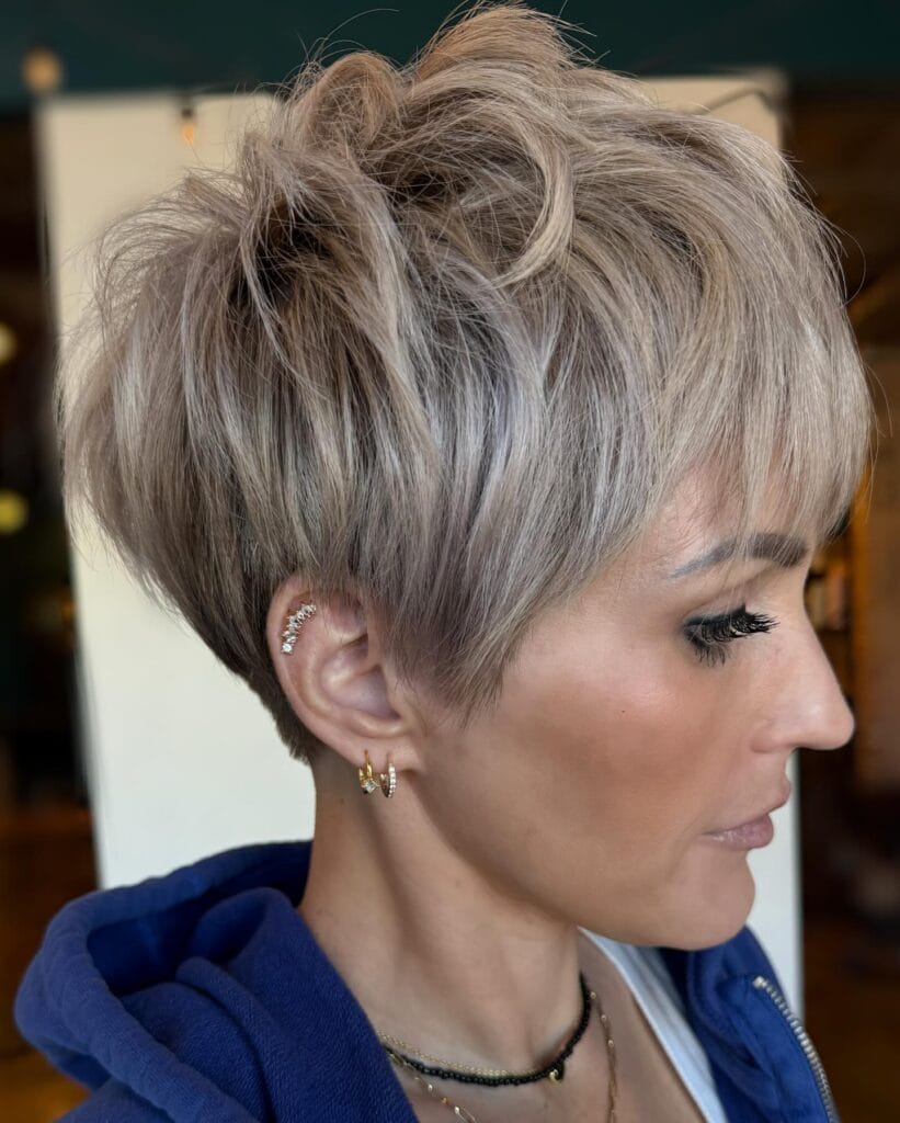 Short textured pixie cut in a cool ash-blonde tone with soft layered volume at the crown, tapered sides, and wispy fringe for a modern, tousled finish.