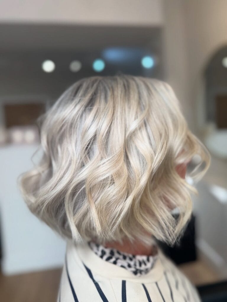 Back view of a shoulder-length blonde bob with soft loose waves, subtle layers, and natural darker roots adding depth to the light platinum color.
