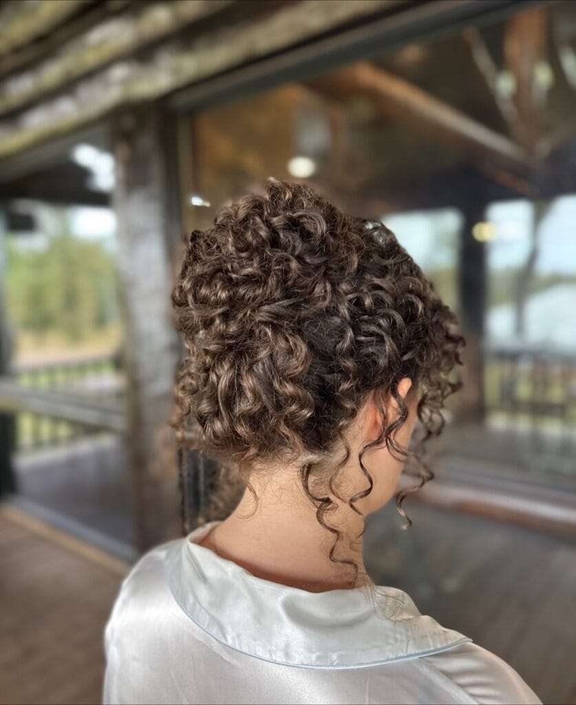 Shoulder-length dark brown curls gathered into a high, voluminous pineapple updo with defined ringlets and a few loose tendrils framing the nape.