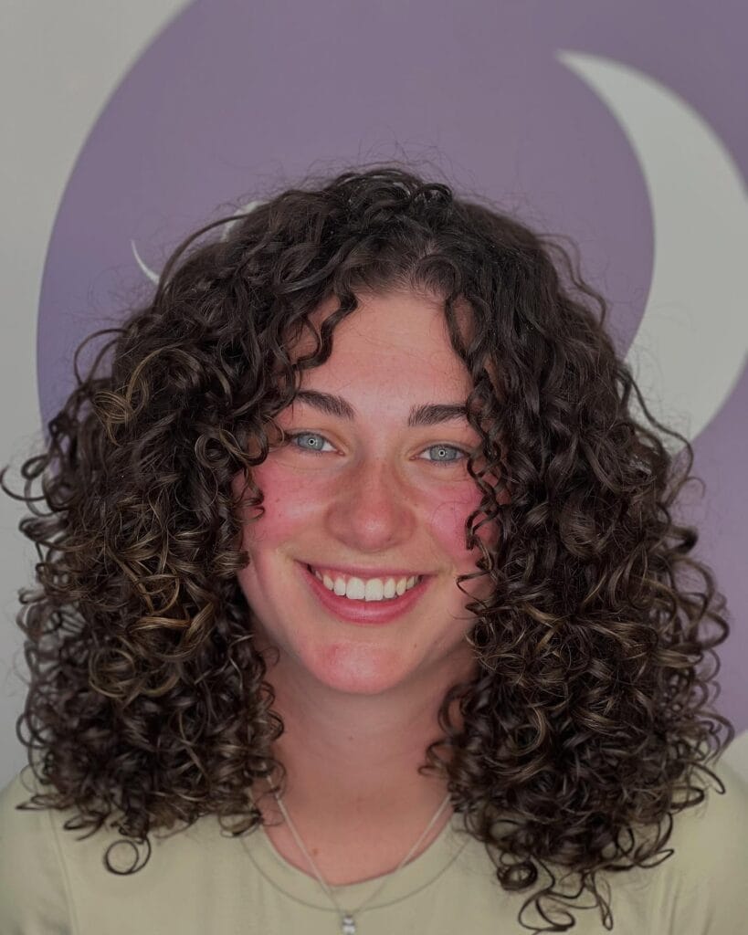 Shoulder-length dark brown curly hair with defined, bouncy ringlets and a soft center part, creating a full, rounded shape around the face.