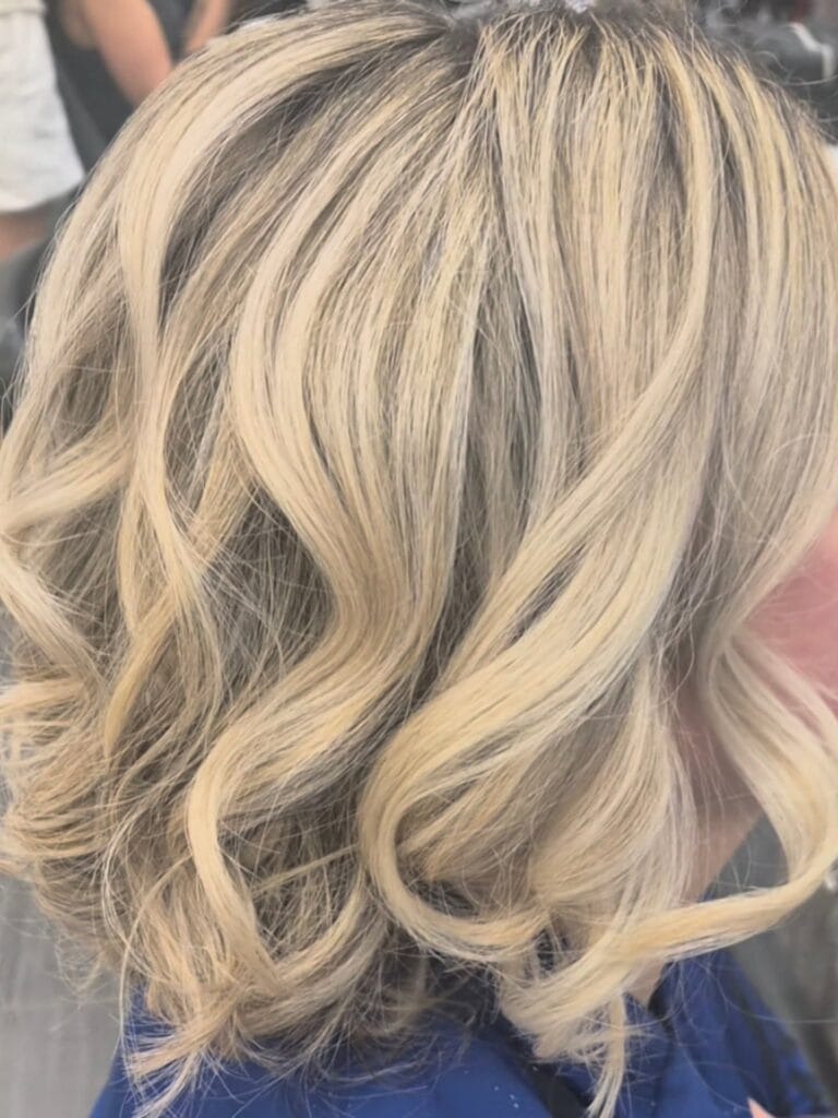 Shoulder-length blonde hair with dark roots styled in loose, soft waves, creating a dimensional balayage look with natural movement.