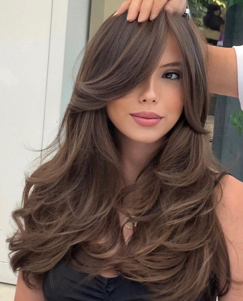 Long, voluminous chestnut-brown hair styled in soft, cascading layers with a deep side part and sweeping bangs that partially cover one eye.