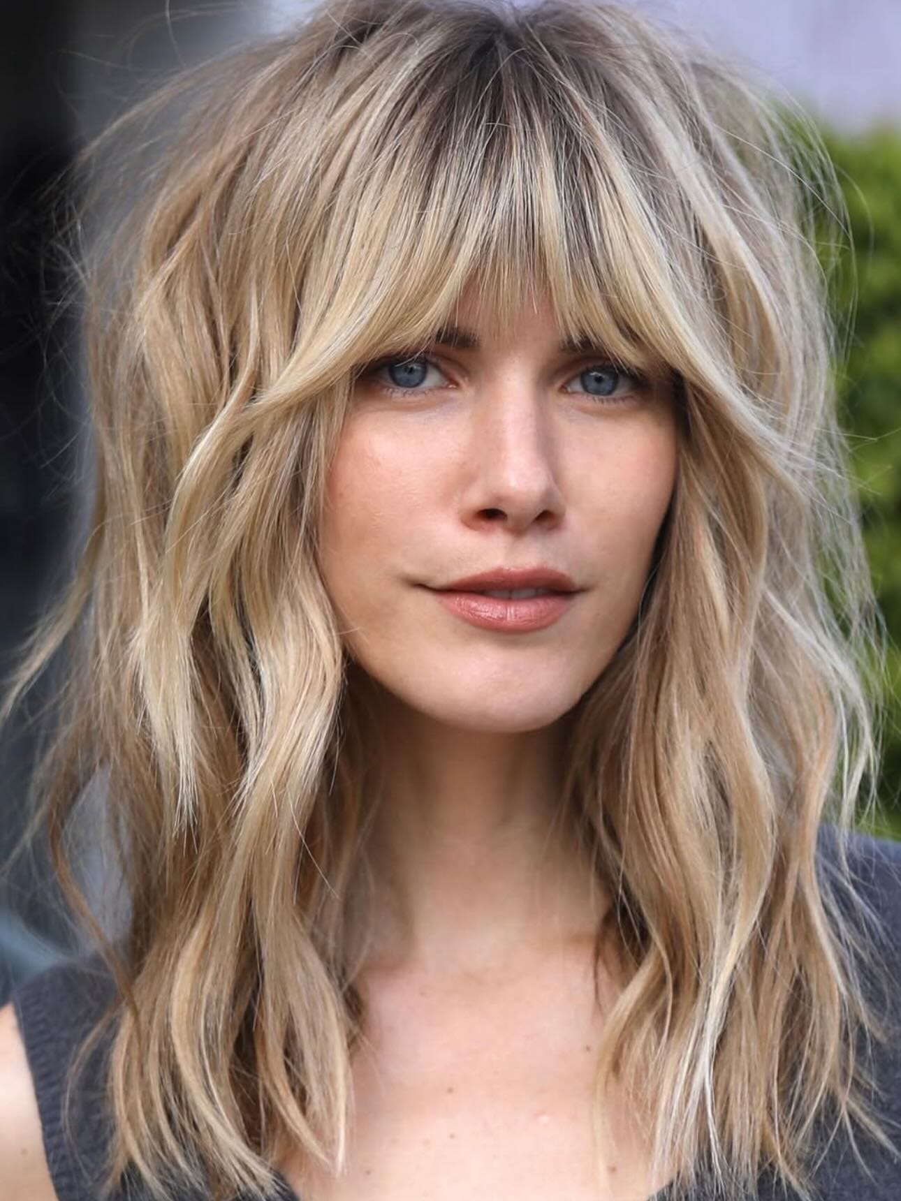 Shoulder-length layered shag haircut with soft wavy texture and wispy curtain bangs that frame the face for a relaxed, modern look.