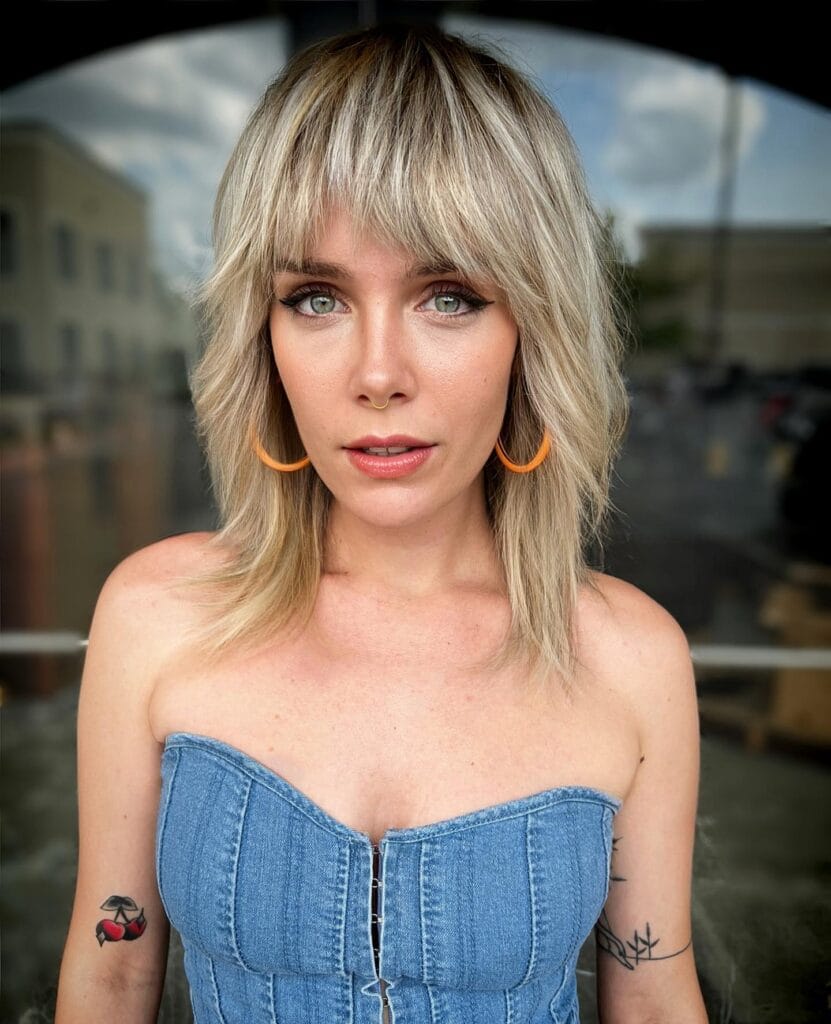 Shoulder-length shag haircut with choppy layers and full textured bangs, styled in soft waves with light blonde tones for a tousled look.