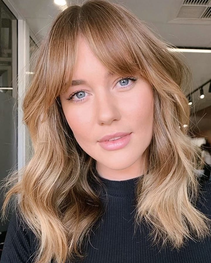 Shoulder-length sandy blonde shag haircut with soft wavy texture, airy layers, and curtain bangs that frame the face for a relaxed, modern look.