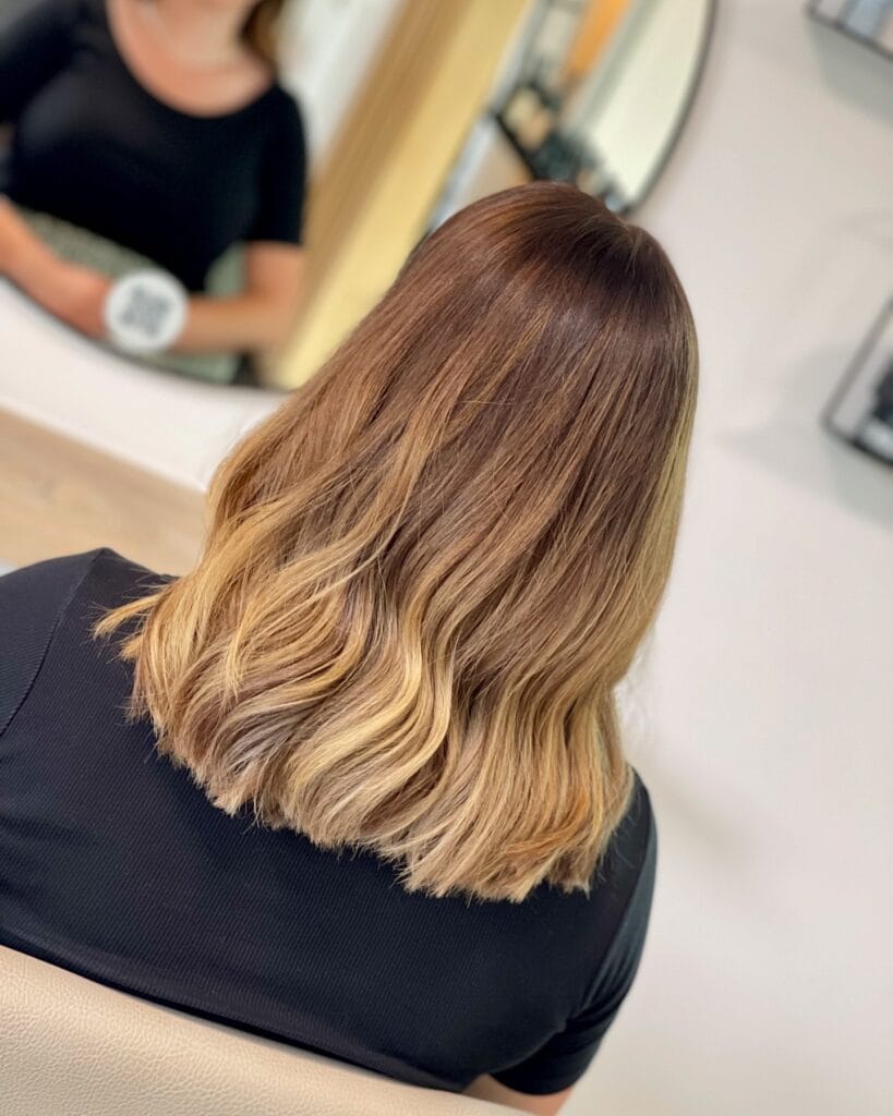 Shoulder-length brunette hair with soft waves, featuring a smooth balayage transition from medium brown roots to warm blonde ends for a natural, dimensional look.
