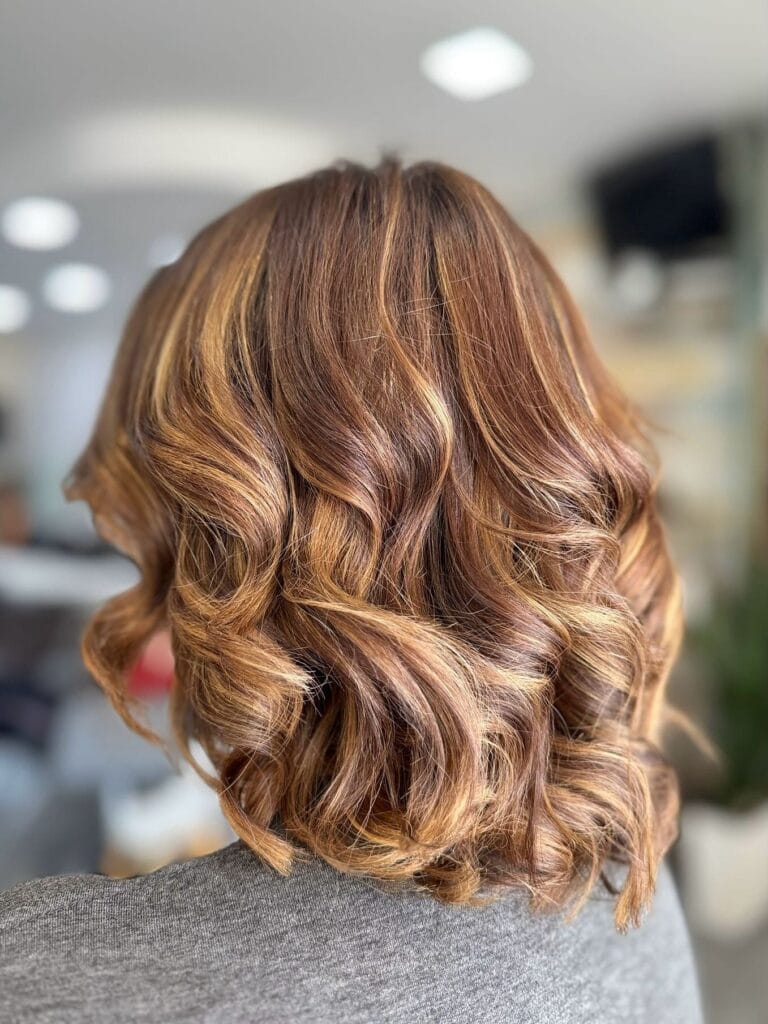 Shoulder-length wavy bob with rich auburn tones and soft caramel highlights that add warmth and dimension throughout the loose curls.
