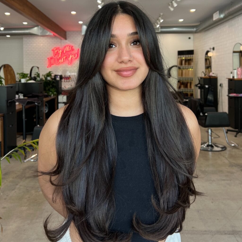 Long, dark brunette hair styled with a center part, cascading in soft, voluminous layers with face-framing pieces and lightly curled ends for a smooth, bouncy finish.