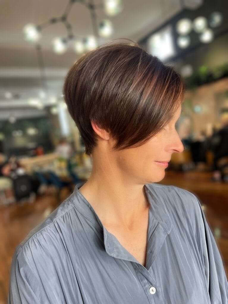 A woman with a sleek brunette bixie cut showing a tapered back and smooth side layers in a side profile view.
