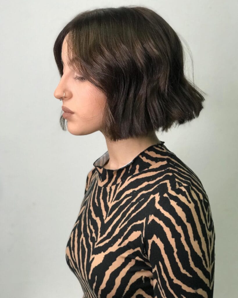 A woman with a chin length dark brunette blunt bob styled in loose waves shows her profile against a light background.