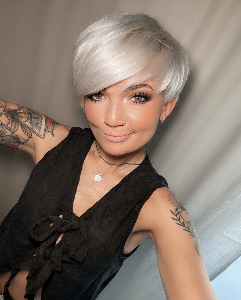 A smiling woman with a sleek icy white pixie cut and long side swept bangs looks at the camera.