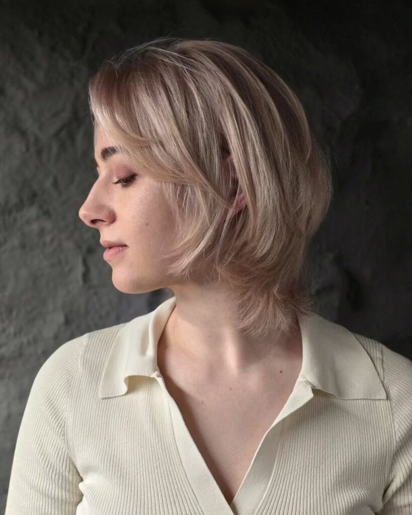 A woman with a feathered rose gold bixie cut and wispy face framing layers is shown from the side.