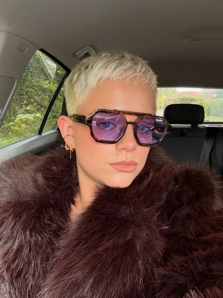 A woman with a very short textured bleach blonde pixie cut wears purple tinted aviator glasses and a faux fur coat.