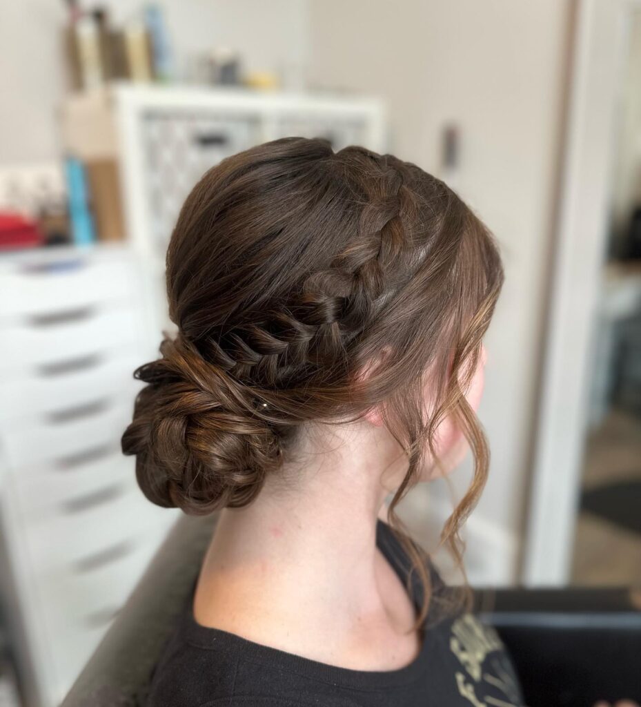 A woman with brunette hair featuring a side Dutch braid that transitions into a textured low knotted bun with loose wavy face framing strands.
