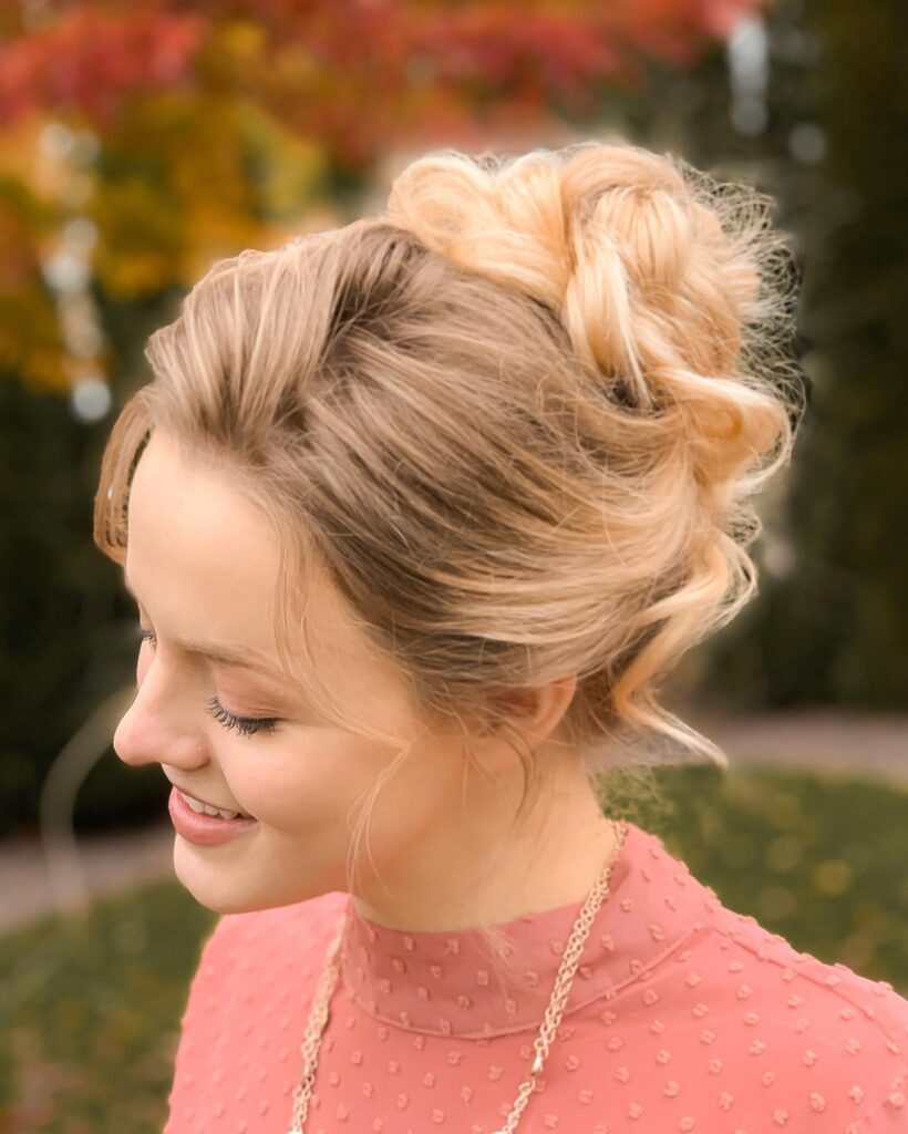 A woman with honey blonde hair styled in a high voluminous braided bun with loose tendrils framing her face.