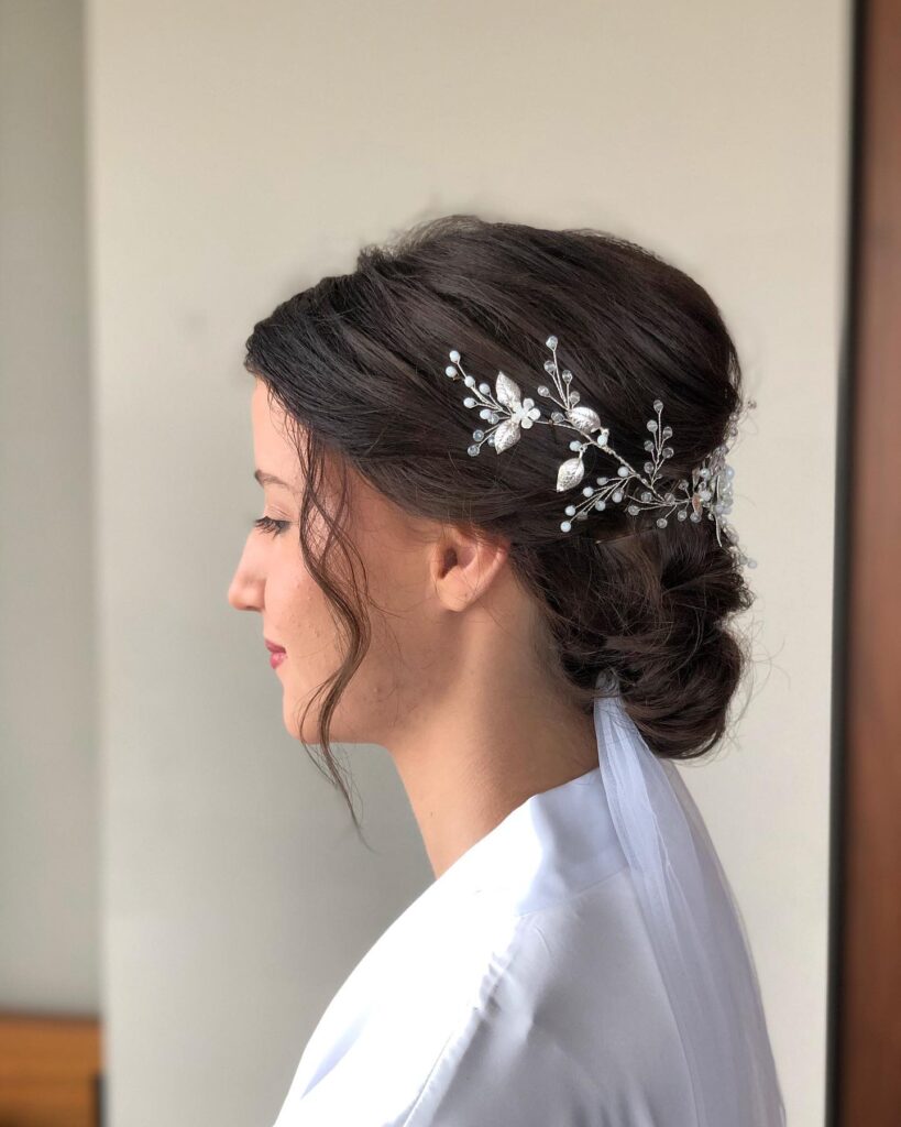 A side profile of a woman with dark brown hair styled in a soft low bun decorated with a silver leafy hair vine and loose wavy strands framing her face.