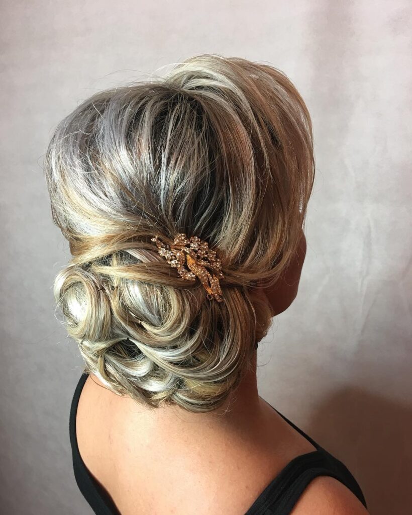 A woman from the back showing an ash blonde low bun with voluminous swept layers and a decorative gold floral hair clip.