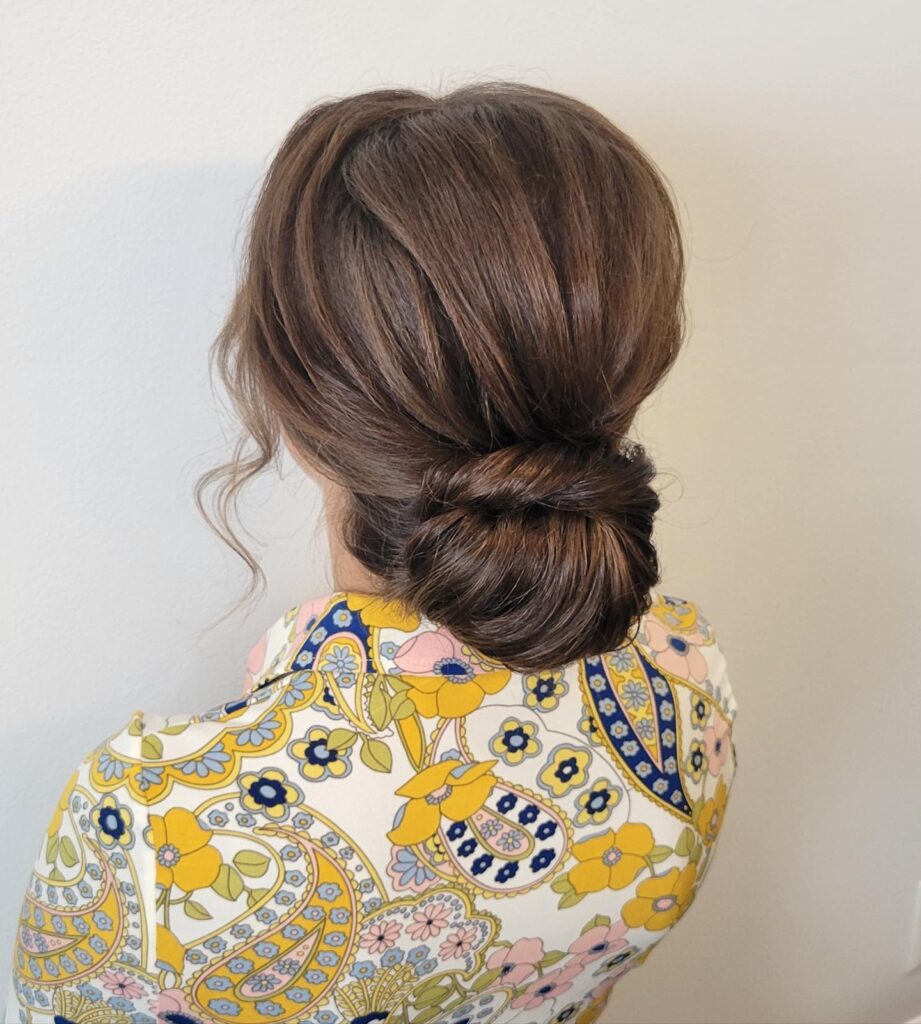 A woman from the back showing a sleek low brunette bun with a twisted wrap around the base and soft wavy strands framing the side.