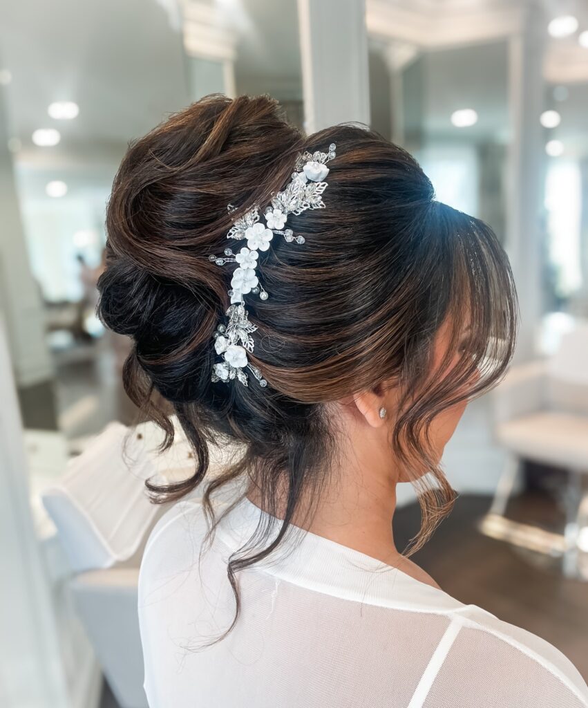 A side view of a woman with dark brown hair styled in a large textured high bun decorated with a white floral hair vine and loose wavy face framing pieces.