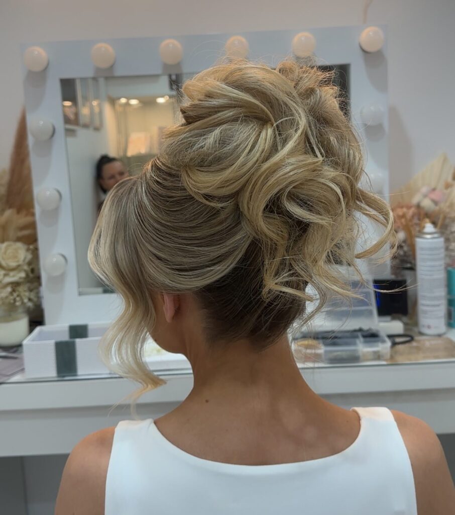 A back view of a woman with multi-tonal blonde hair styled in a high, voluminous updo featuring prominent looped waves and loose face-framing curls.