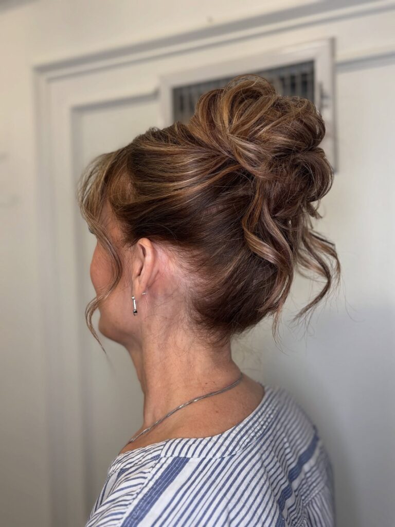 A woman from the side showing a voluminous high honey brown messy bun with curled sections, blonde highlights, sweeping side bangs, and loose tendrils framing her face.