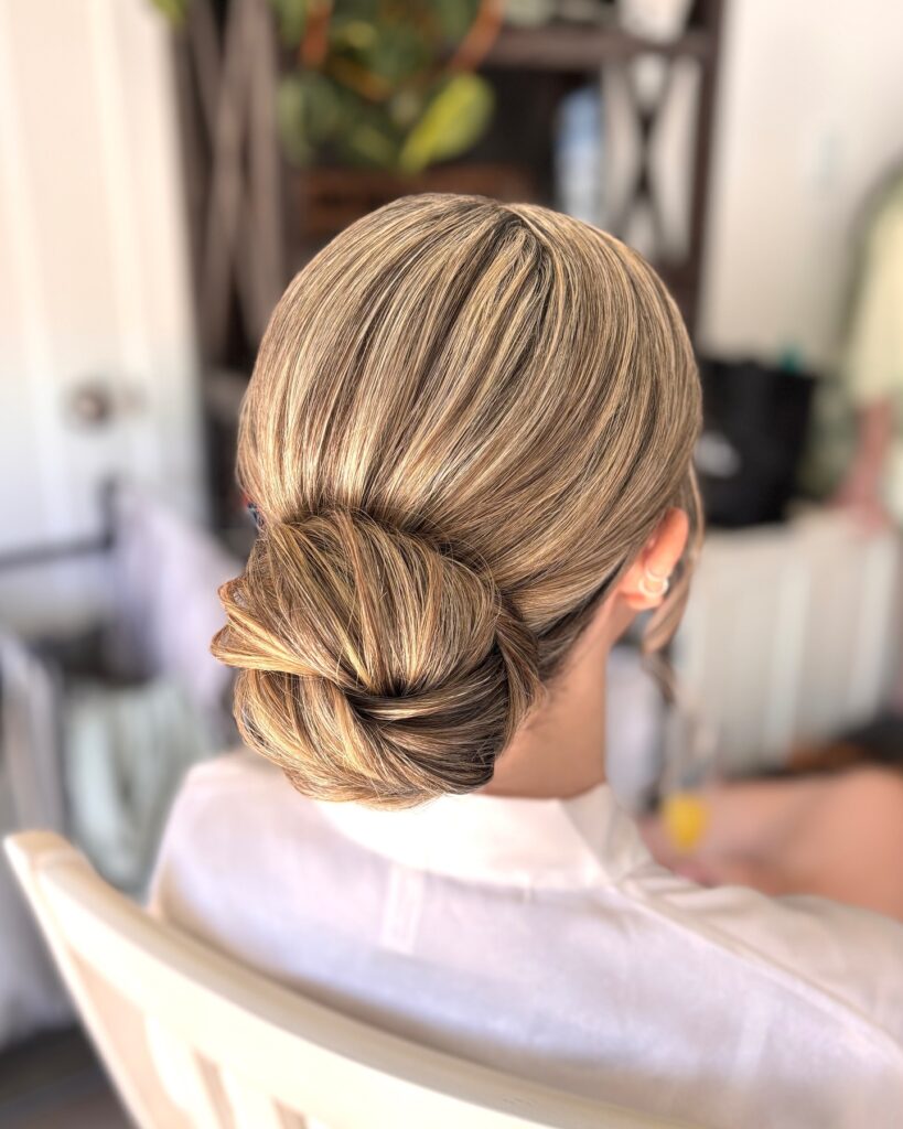 A back view of a woman with highlighted blonde and brown hair styled into a perfectly sleek and smooth low bun at the nape of her neck.