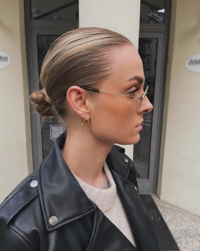 A side profile of a woman wearing glasses with blonde hair styled in a sleek wet look low micro bun and a black leather jacket.