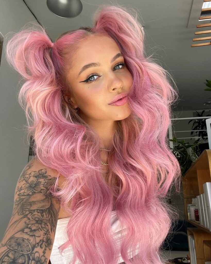 A woman with long wavy pastel pink hair styled into two high pigtails with loose voluminous layers.