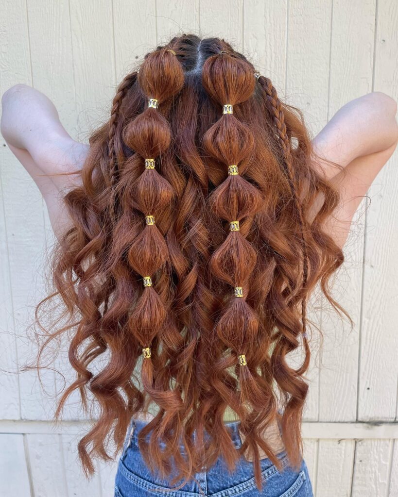 A woman with long curly copper hair wears two central bubble braids decorated with gold metallic rings.