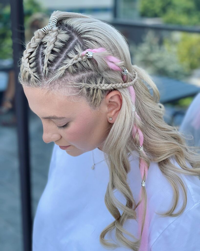 A woman with intricate braided top sections in her platinum blonde hair, featuring multiple small silver wire coils and beads and a pink yarn braid detail on one side.