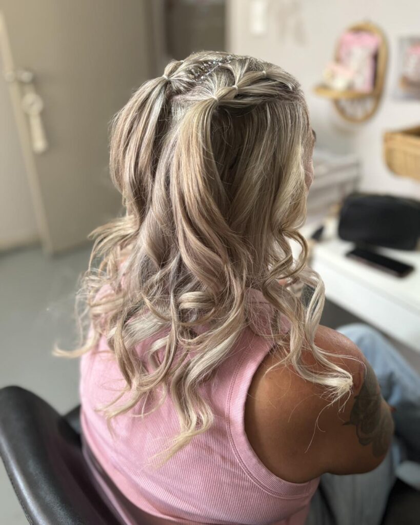 A woman with medium-length blonde hair from behind, wearing it in a intricate criss-crossed half up ponytail with loose curls and a line of rhinestones on her center part.