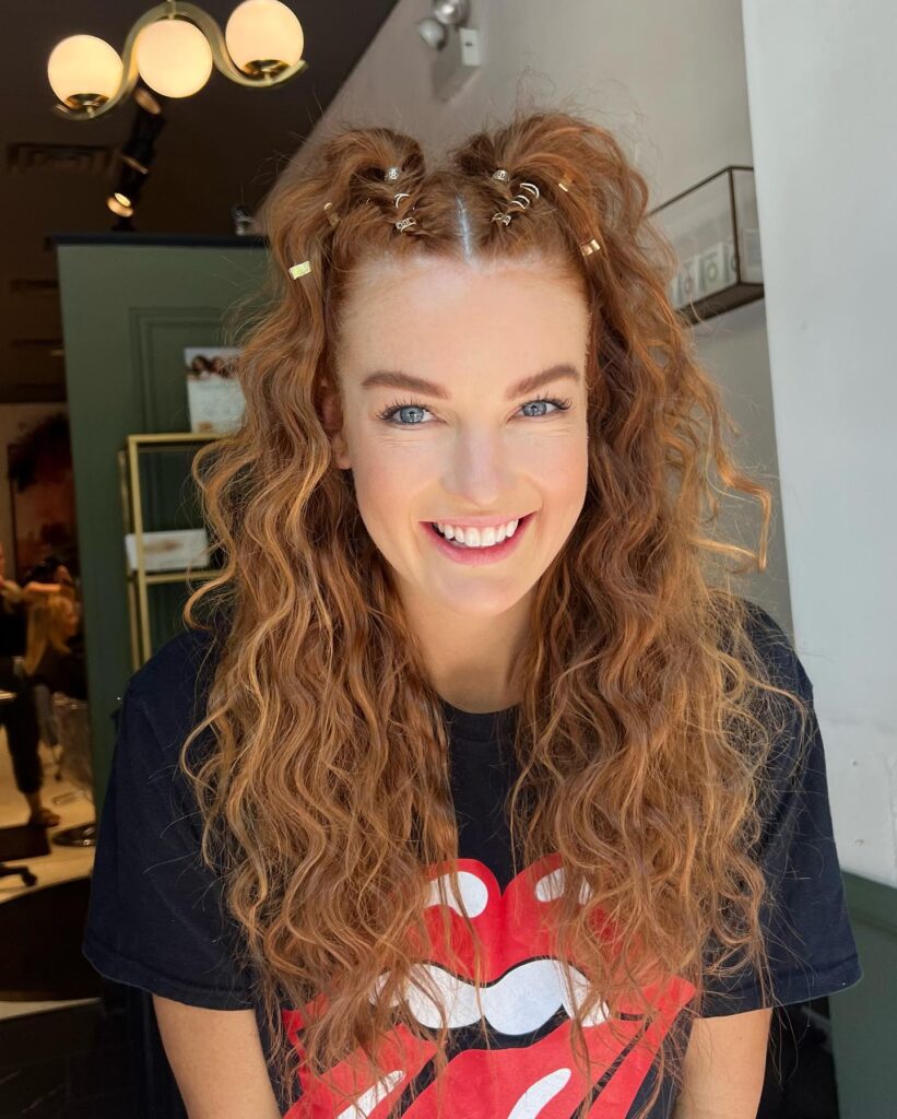 A smiling woman with long curly copper hair wearing two high half-up ponytails decorated with small gold metallic hair rings.