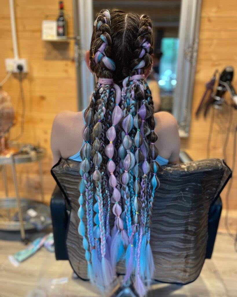 A rear view of a person with long dark hair integrated with blue and purple extensions styled into Dutch braids, bubble sections, and small braids.