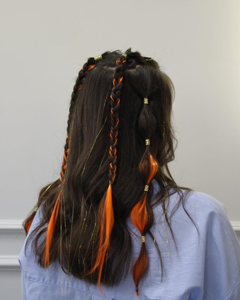 A woman with long dark wavy hair featuring two braids and a bubble ponytail accented with bright orange extensions and gold tinsel.