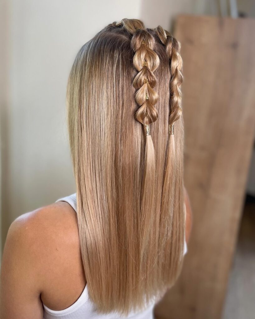 A woman with long straight blonde hair styled with two small pull through braids on top secured by gold decorative beads.