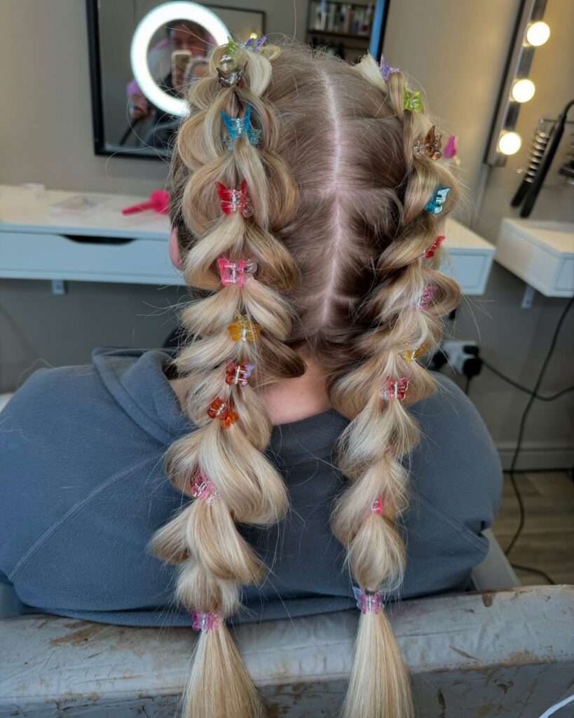 A rear view of a woman with long blonde hair styled into two bubble braids with colorful butterfly-shaped clips.