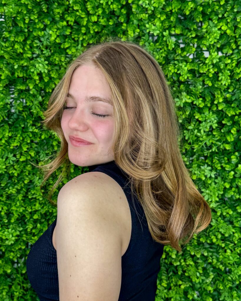 A woman with a shoulder length honey blonde butterfly haircut featuring golden highlights and face framing layers.