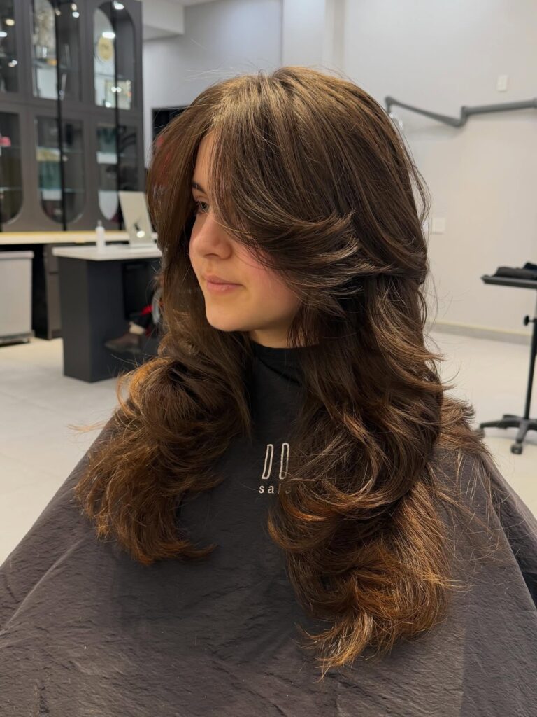 A woman with long honey brown hair featuring voluminous butterfly layers and styled face framing curtain bangs.