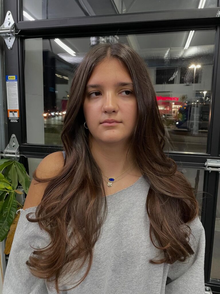 A young woman with long chocolate brown hair featuring soft layers and a middle part styled with gentle waves at the ends.