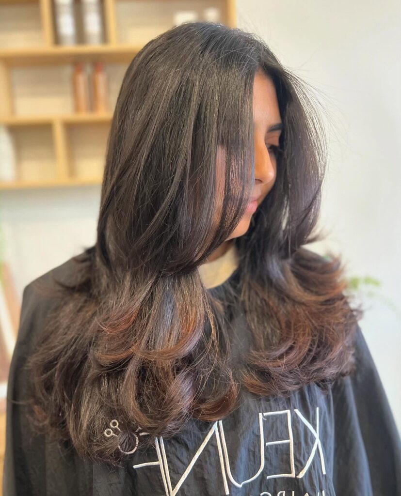 A woman with long dark brown hair styled in a voluminous blowout with face framing layers and feathered ends.