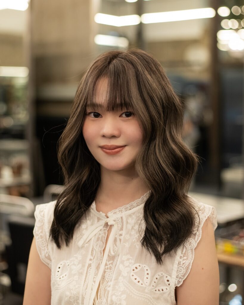 A young woman with a dimensional mushroom brown wavy lob featuring thin see through bangs styled in loose curls.